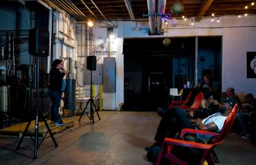 A comedian performing on stage to a small audience in an industrial-style venue.