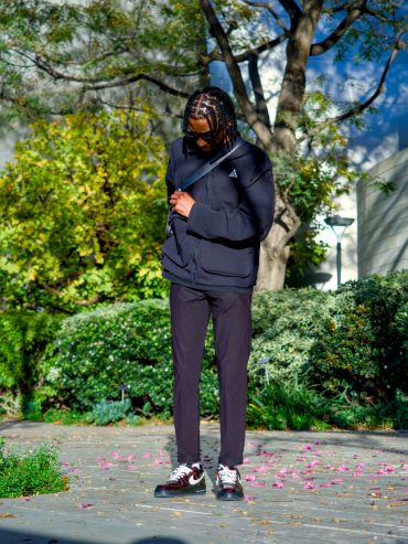 A man in black streetwear stands on a path surrounded by greenery.