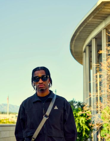 A man wearing sunglasses and a black jacket stands outdoors near a modern curved building.