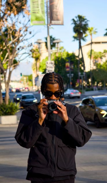 Young man with braided hair taking a photo with a camera on a sunny city street.