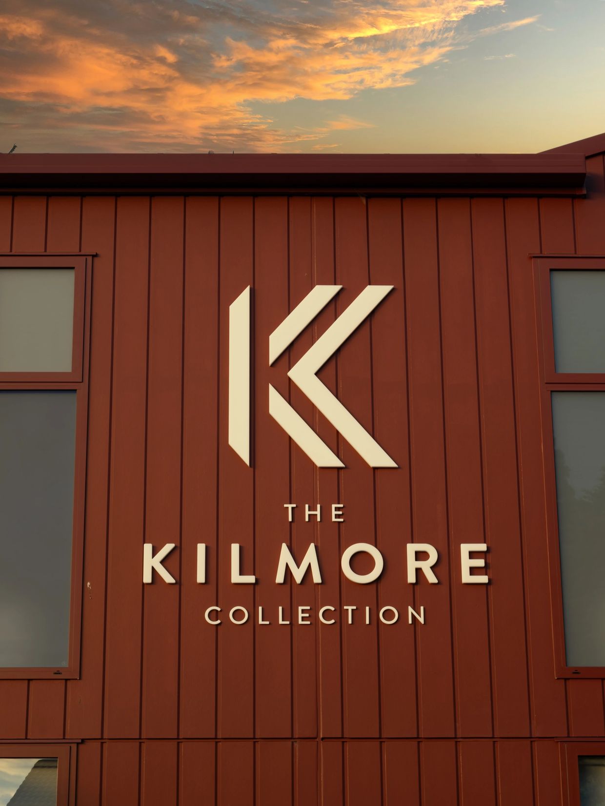Sign for The Kilmore Collection on a red building under a colorful sunset sky.