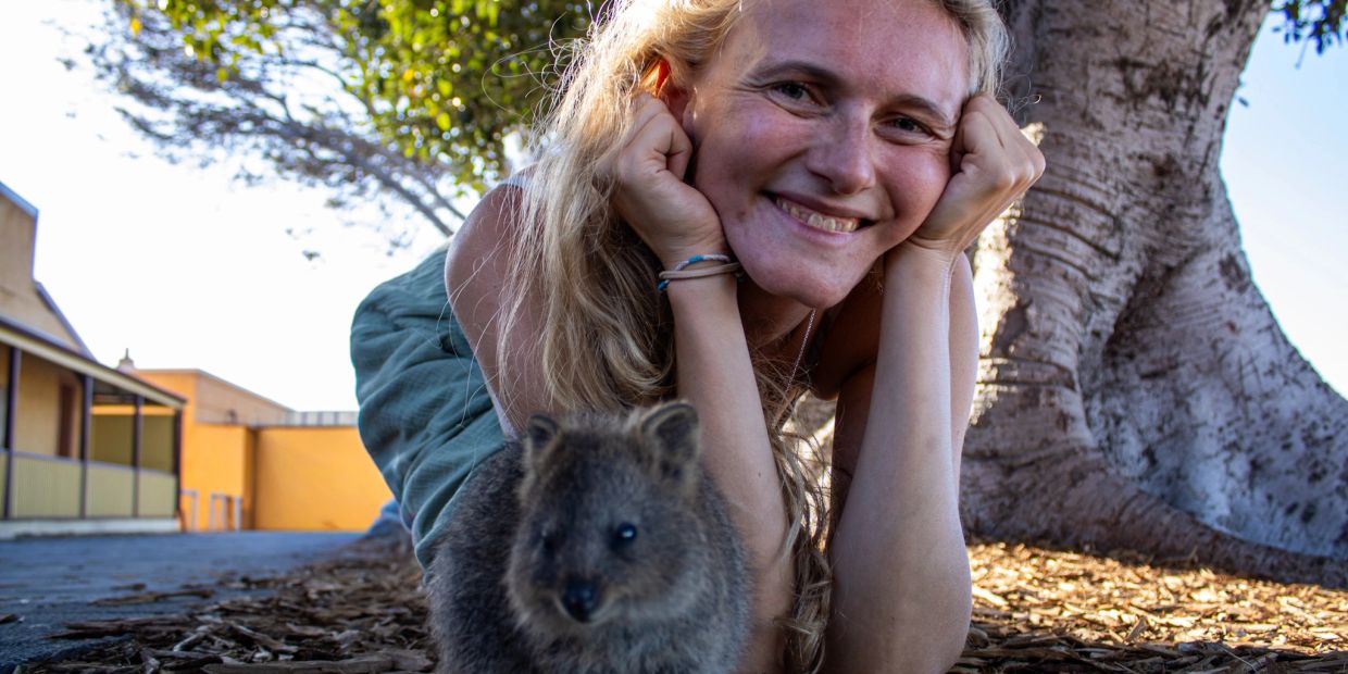 Frau posiert lächelnd neben einem Quokka unter einem Baum.