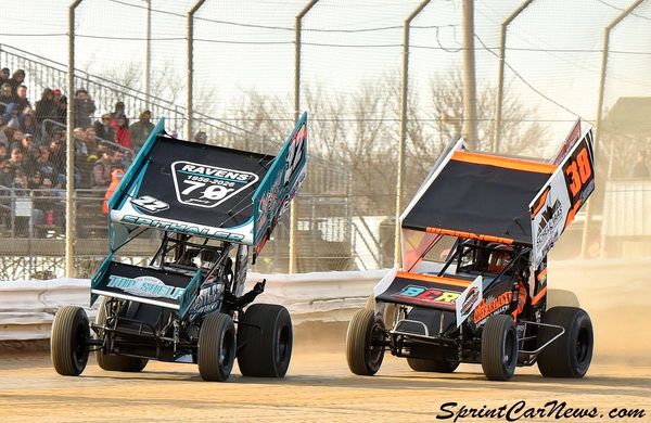 Two sprint cars racing on a dirt track with spectators in the background.