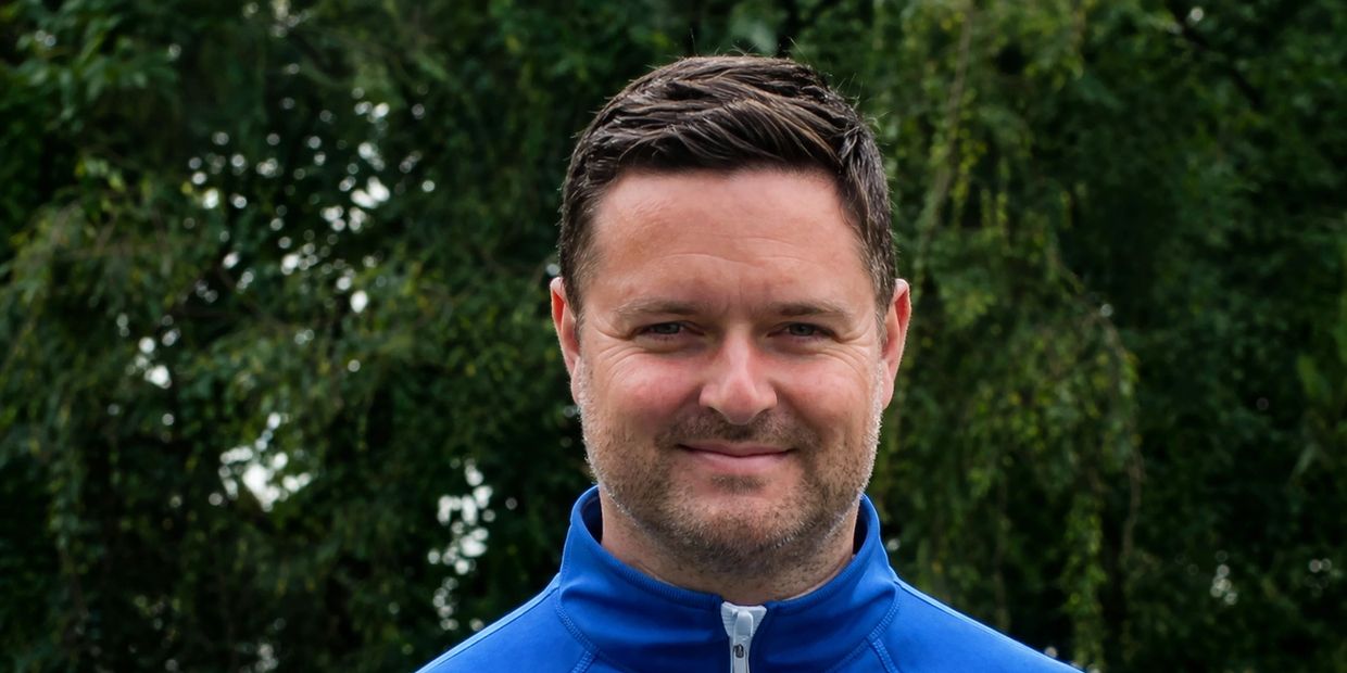 Man with short dark hair wearing a blue jacket smiling outdoors.