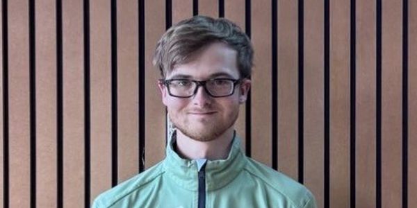 Man with glasses and a light green jacket in front of a wooden panel wall.