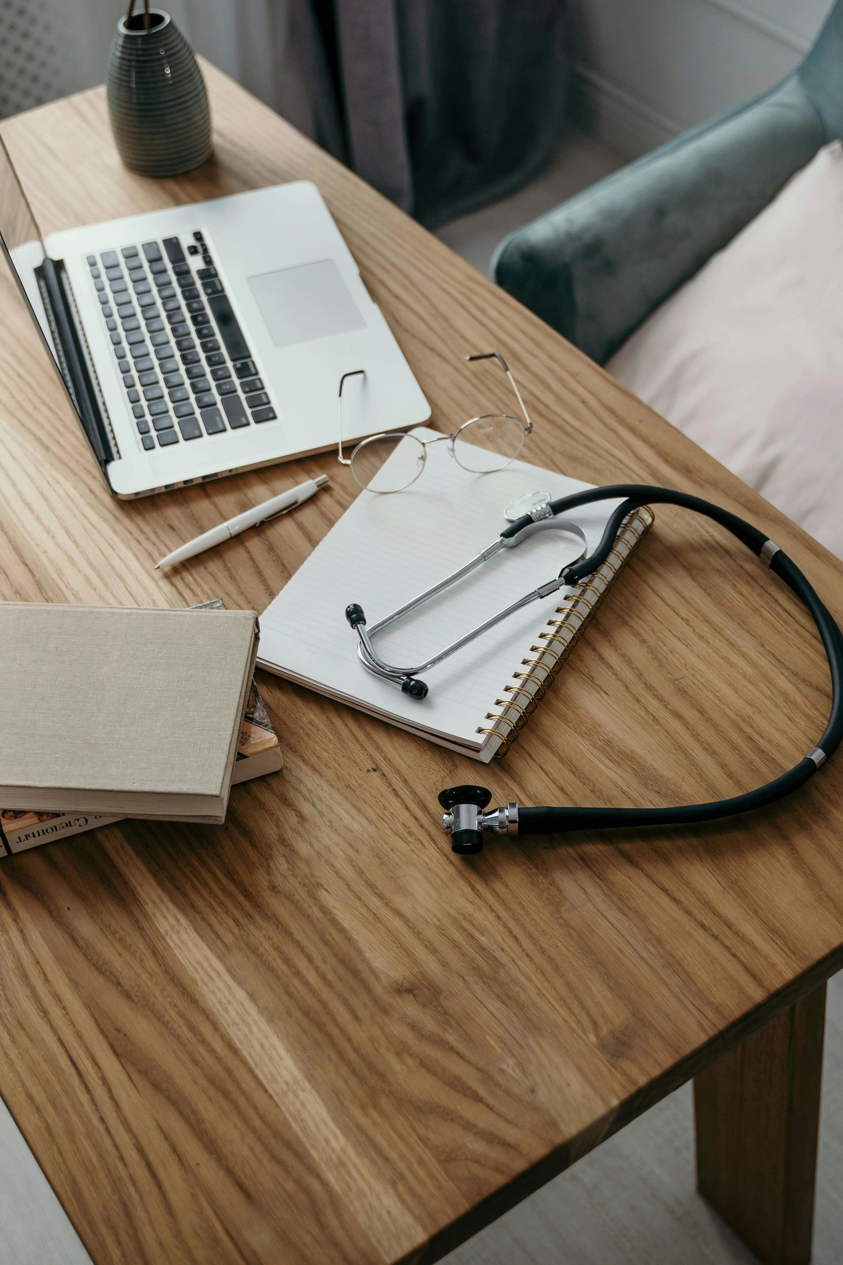 A doctor's desk with a computer, books, a pair of glasses, a pen, a blue vase, and a stethoscope. 