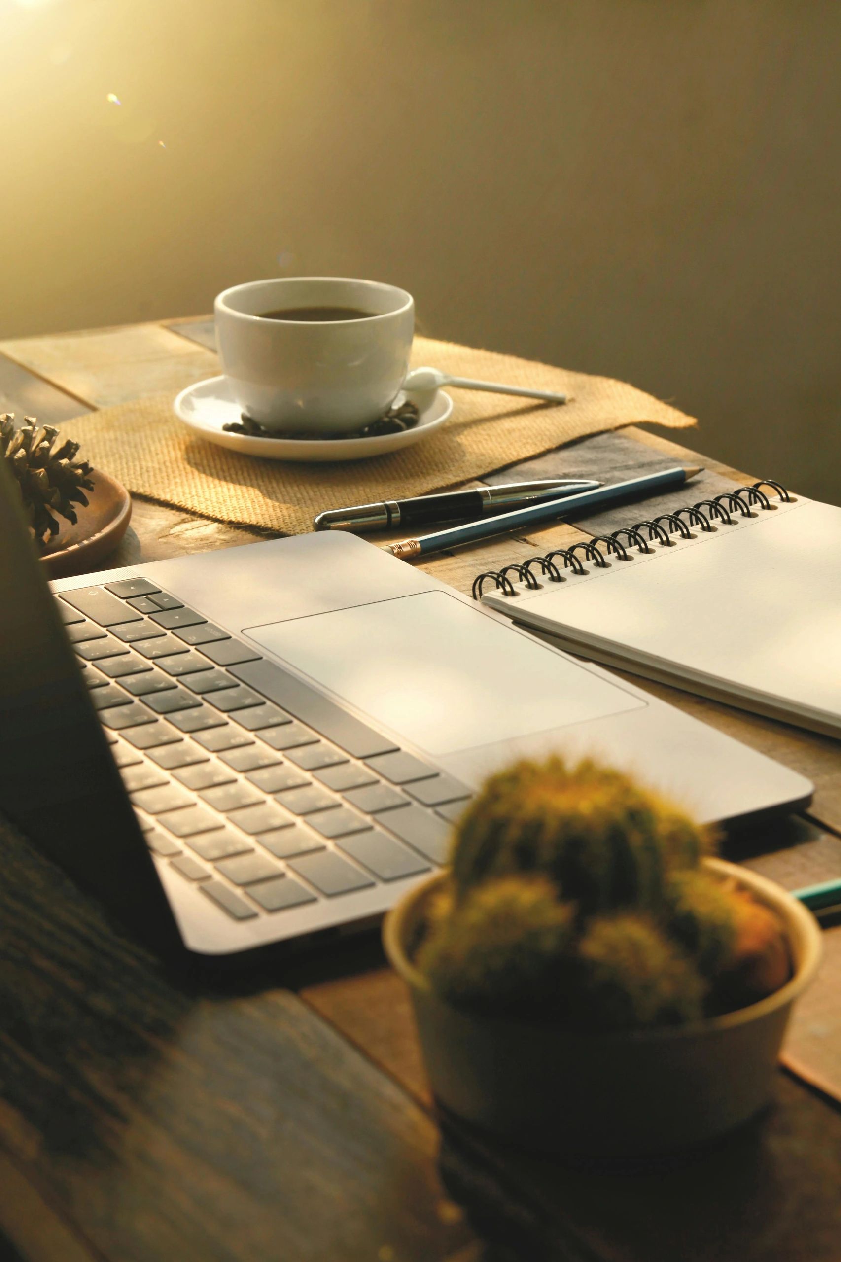 Warm morning workspace with coffee, laptop, notebook, and pens.