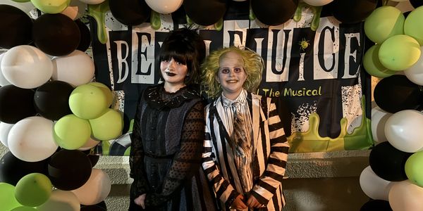 Two kids dressed as Beetlejuice characters stand in front of a themed balloon arch.