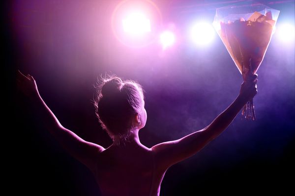 Performer holding flowers on stage, arms raised to bright lights.