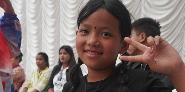 Smiling girl making a peace sign with people sitting in the background.
