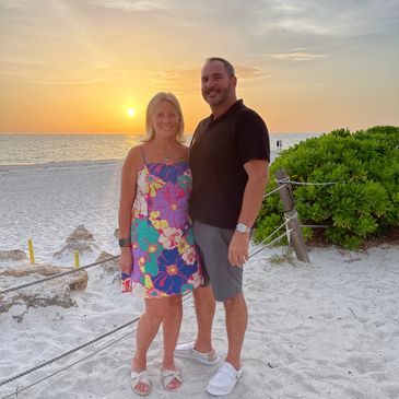 Couple posing on the beach at sunset with ocean and greenery in the background.