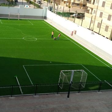 A green soccer field with two people near the sideline in a residential area.