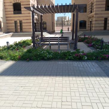Wooden pergola with benches in a courtyard surrounded by flowers and buildings.