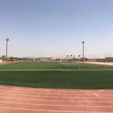 Empty sports field with green turf and red running track under clear sky.