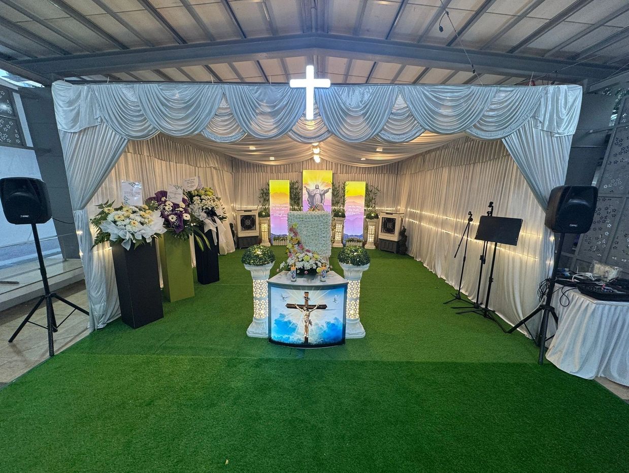A solemn Christian memorial setup with flowers, a crucifix, and white drapery under a roof.