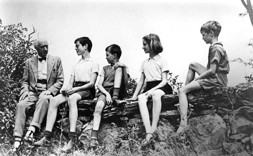 An elderly man with four children sitting on a log outdoors.