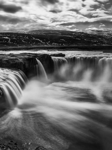 Black and white photo of a wide waterfall flowing over rocky cliffs.