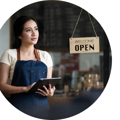 Woman in apron holding tablet stands near an open sign in a store.