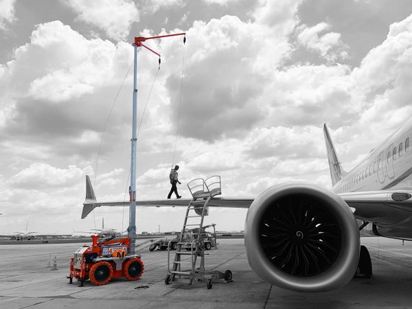Person walking on airplane wing with crane and ladder nearby under a cloudy sky.