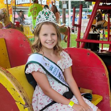 Young girl wearing a crown and sash, smiling at a fair.