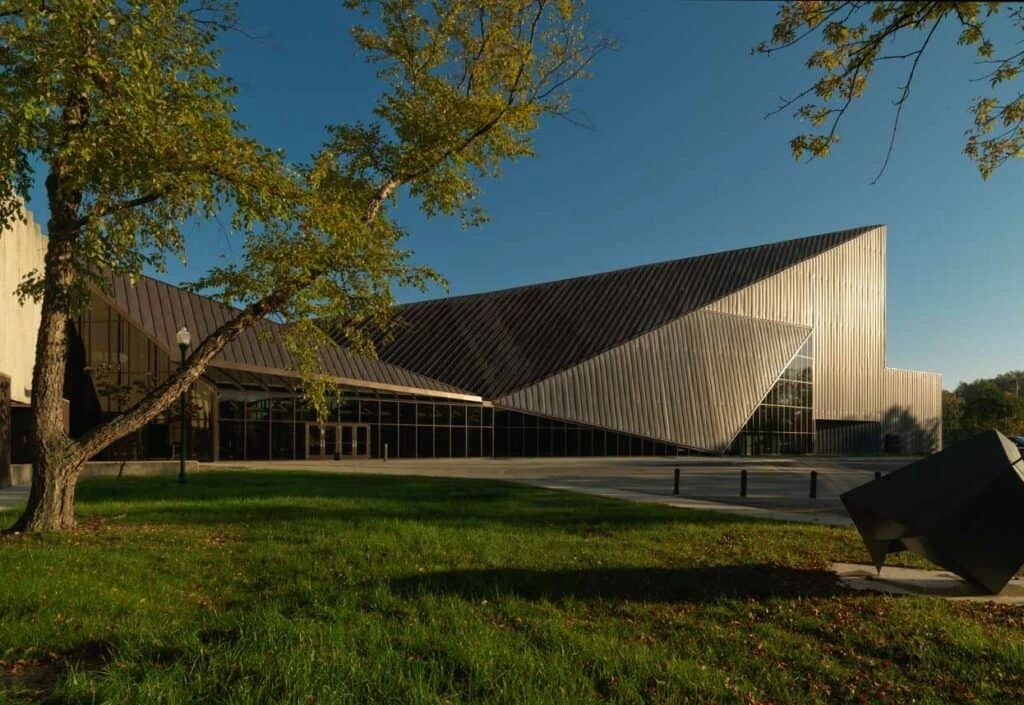 A modern building with a unique angular design and large glass windows, surrounded by a grassy area with a tree and a sculpture.