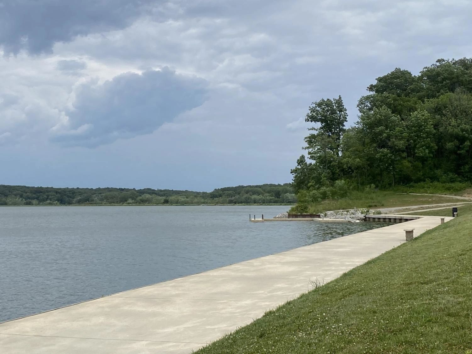 A concrete dock extends into a lake, surrounded by grass and trees under a cloudy sky.