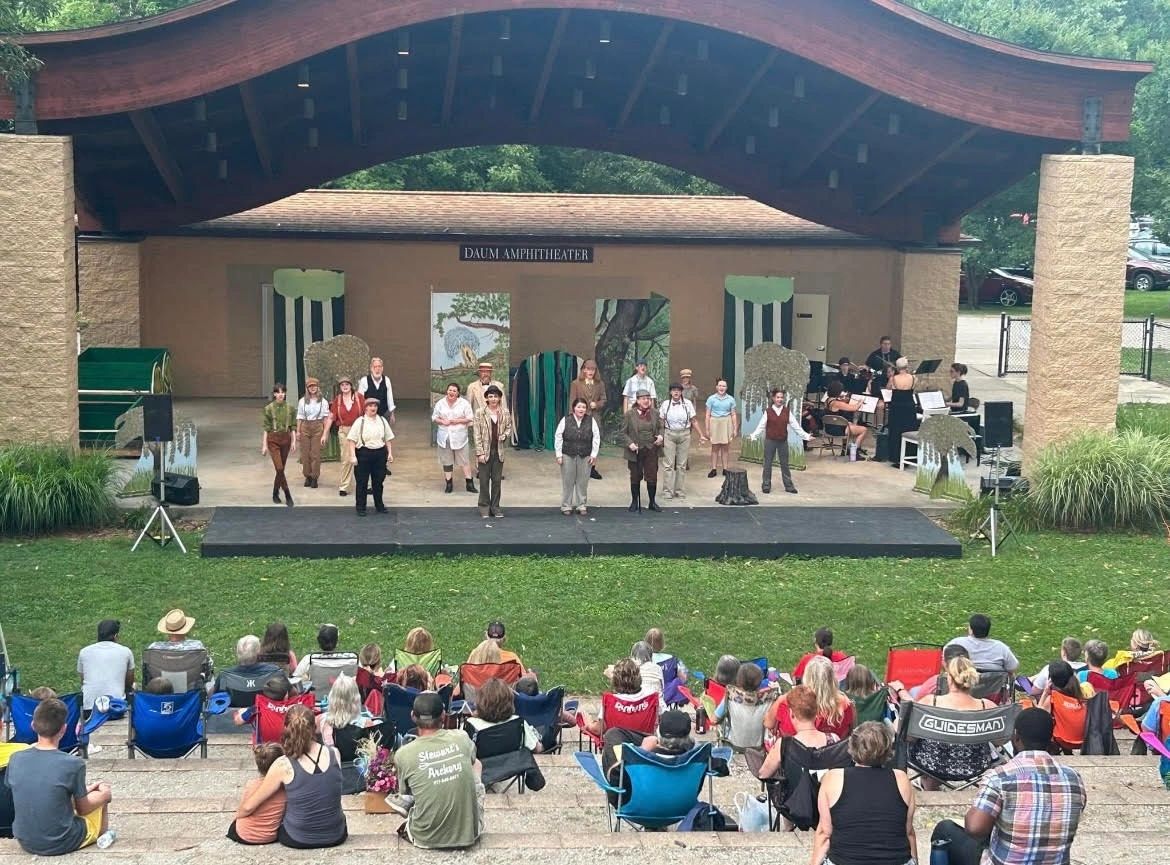 A group of people on stage in an outdoor amphitheater with a roof, with an audience seated in front of them.