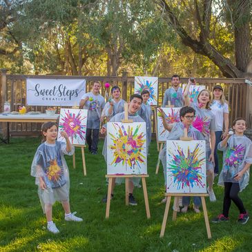 Children proudly display colorful splatter paintings at an outdoor art event.