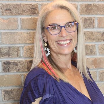 Smiling woman with glasses and colorful hair streaks in front of a brick wall.