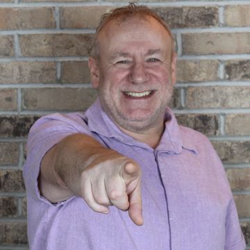 Smiling man in a purple shirt pointing forward.