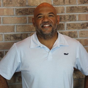 Man smiling, standing against a brick wall, wearing a light blue polo shirt.