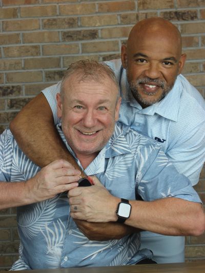 Two men smiling, one hugging the other from behind, both wearing watches.