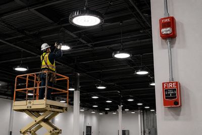 Worker on lift installing or inspecting ceiling lights in a large industrial space.