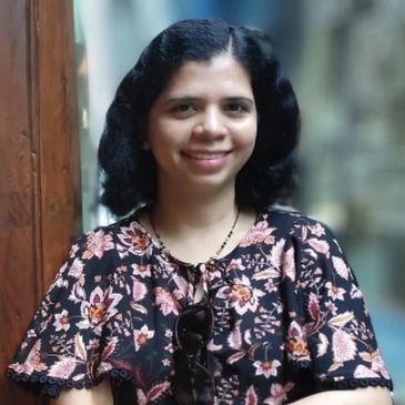 Woman smiling with arms crossed, wearing a floral dress and standing by a wooden door.