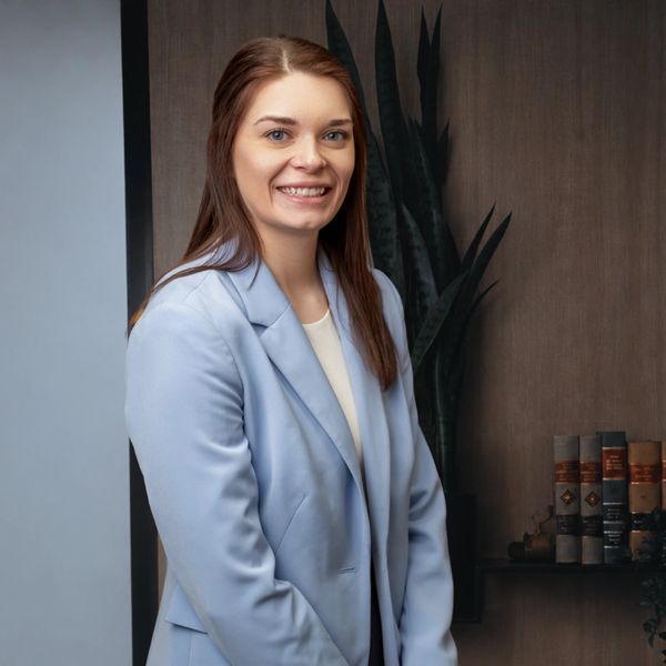 Smiling woman in a light blue blazer standing indoors with books and a plant in the background.