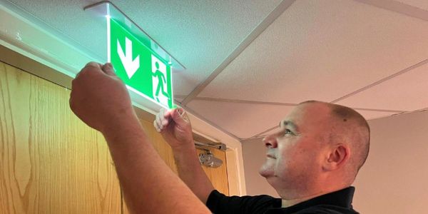 Man installing or adjusting an illuminated emergency exit sign above a door.