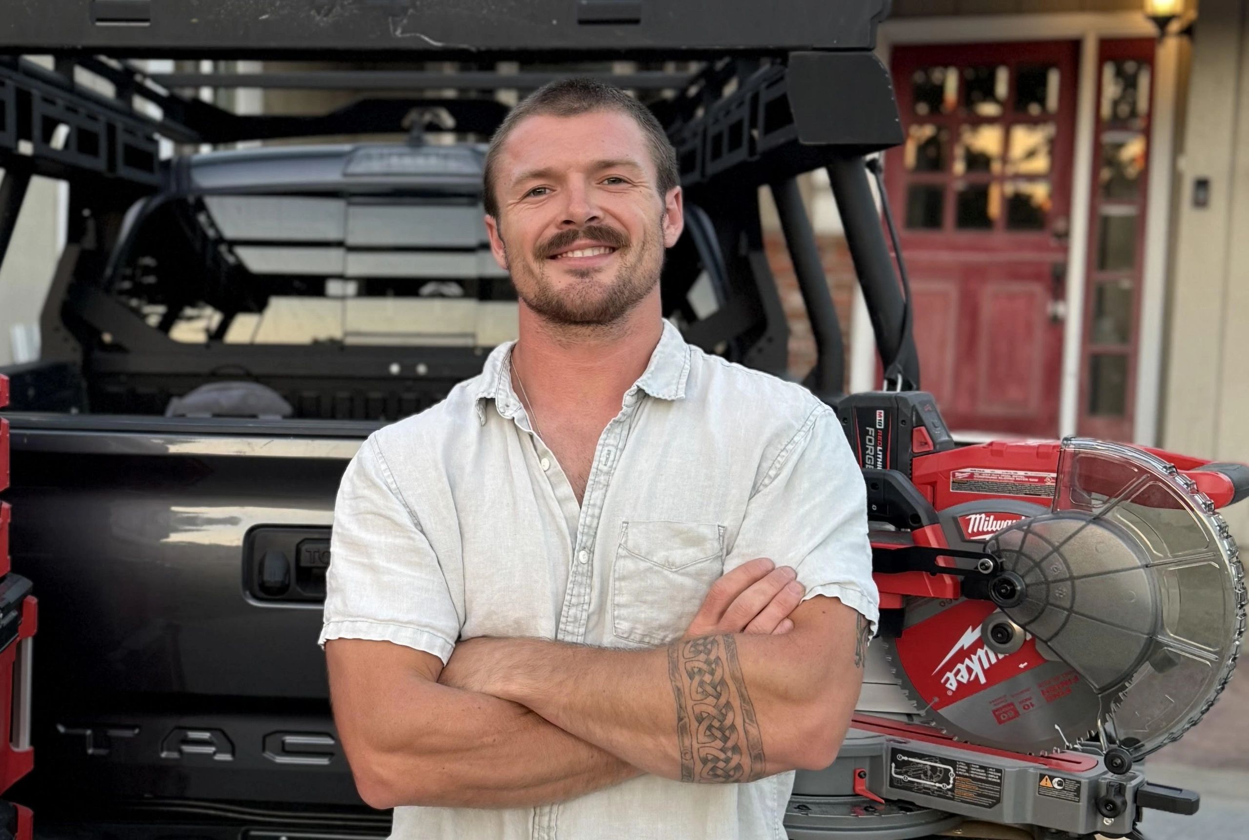 Man with a tattooed arm stands confidently in front of a truck and power tool.