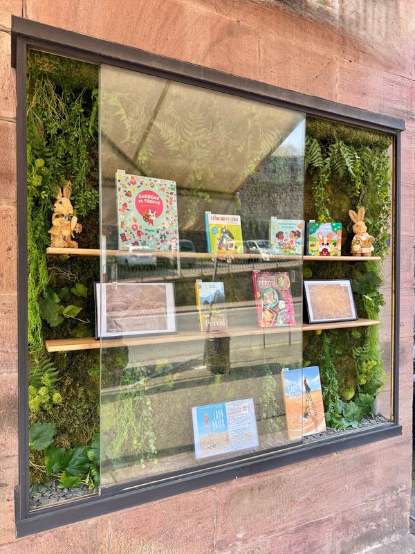 Display case with books, framed pictures, and stuffed rabbits, surrounded by green foliage.