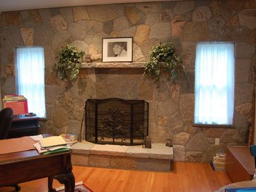 Interior stone fireplace with a mantel, windows, and potted plants.
