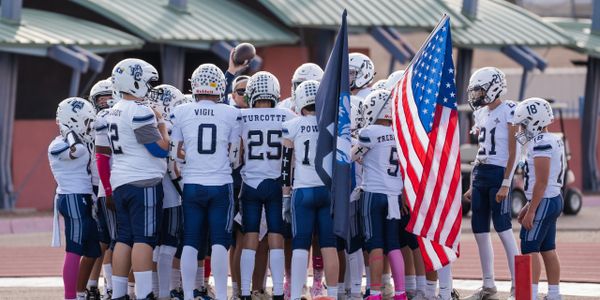 High school football team huddles on field with American flag.