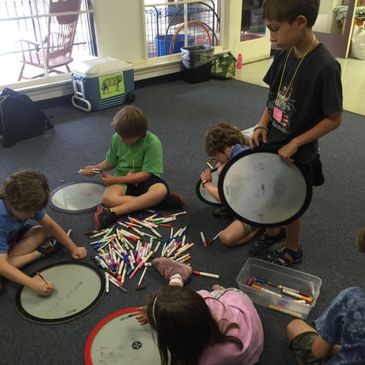 Children sitting on the floor drawing on drum heads with markers.