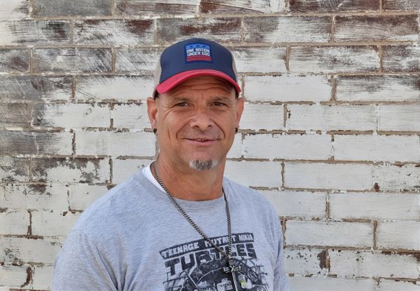 Man wearing a cap and graphic tee in front of a brick wall.