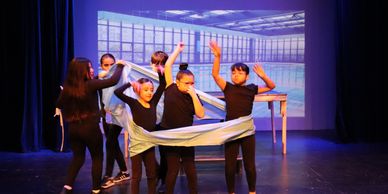 Children performing a play on stage with a backdrop of a modern room.