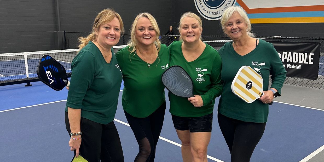 Four women in matching green tops pose on an indoor pickleball court holding paddles.