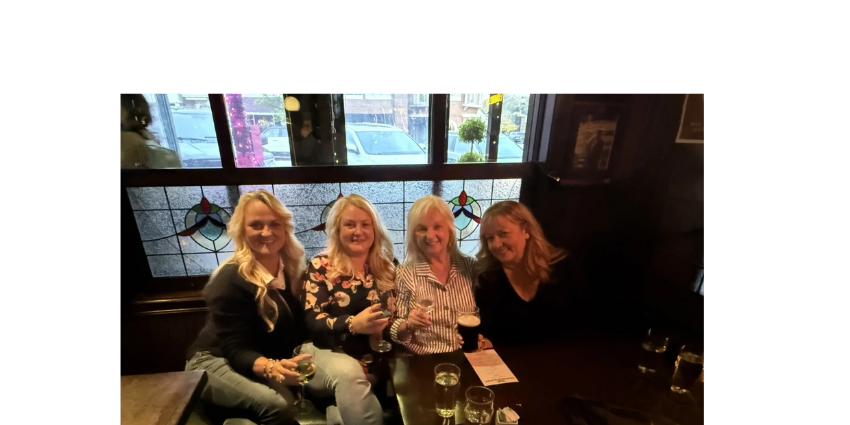 Four women enjoying drinks together at a cozy pub with stained glass windows.