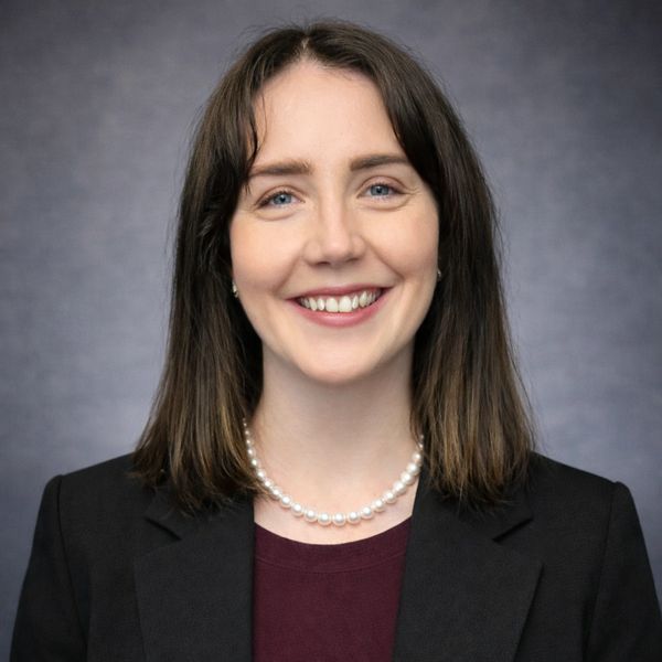 A young woman with brown hair smiles warmly, wearing a black blazer and pearl necklace.
