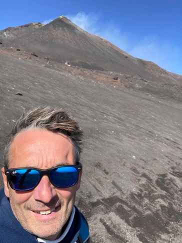 Man in sunglasses taking a selfie on a volcanic mountain with smoke rising.