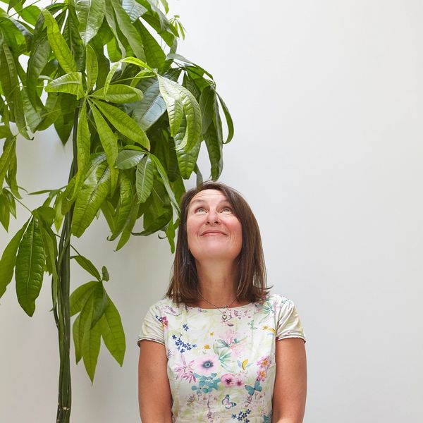 Woman in floral dress looking up beside a tall green plant indoors.