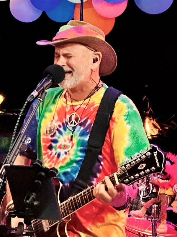 Musician in a colorful tie-dye shirt and hat passionately singing and playing guitar.