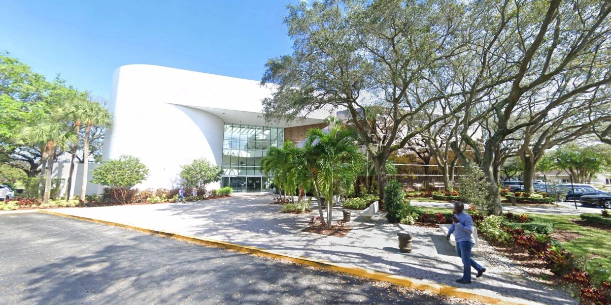 Modern white building surrounded by lush trees and shrubs under a clear blue sky.